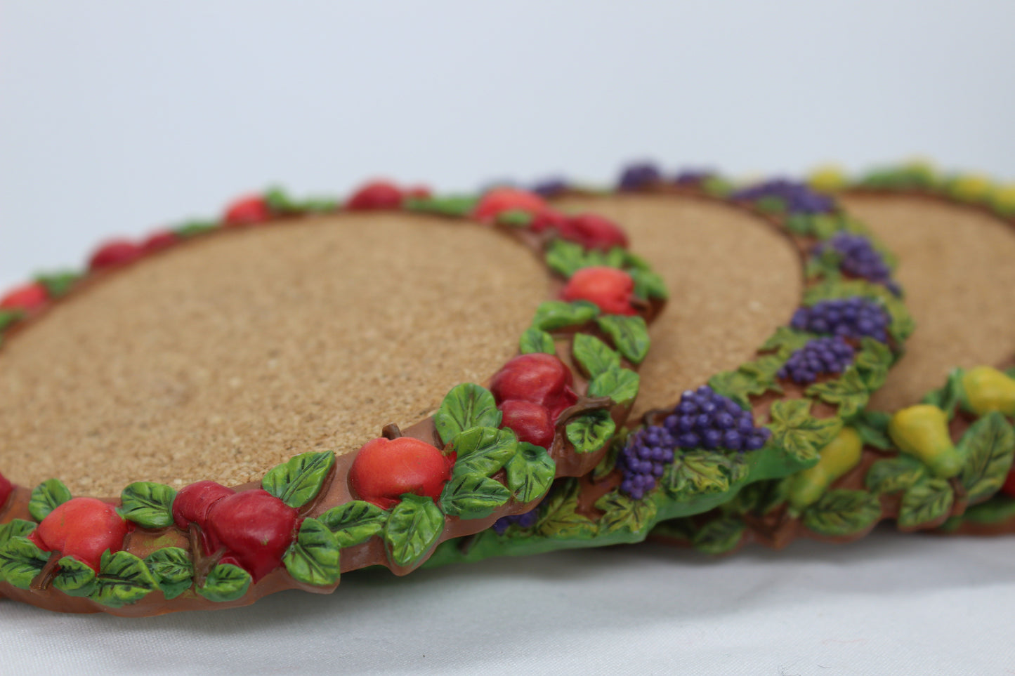 Set of 4 Coasters -  Apple Pattern (2), Grape   Pattern (1); Pear Pattern (1)  Russ Wine Cellar - Berrie and Company  Approx:  3.75” Round  Ceramic With Cork Inlay