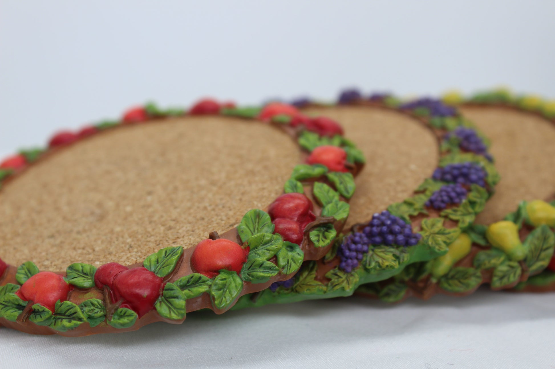 Set of 4 Coasters -  Apple Pattern (2), Grape   Pattern (1); Pear Pattern (1)  Russ Wine Cellar - Berrie and Company  Approx:  3.75” Round  Ceramic With Cork Inlay